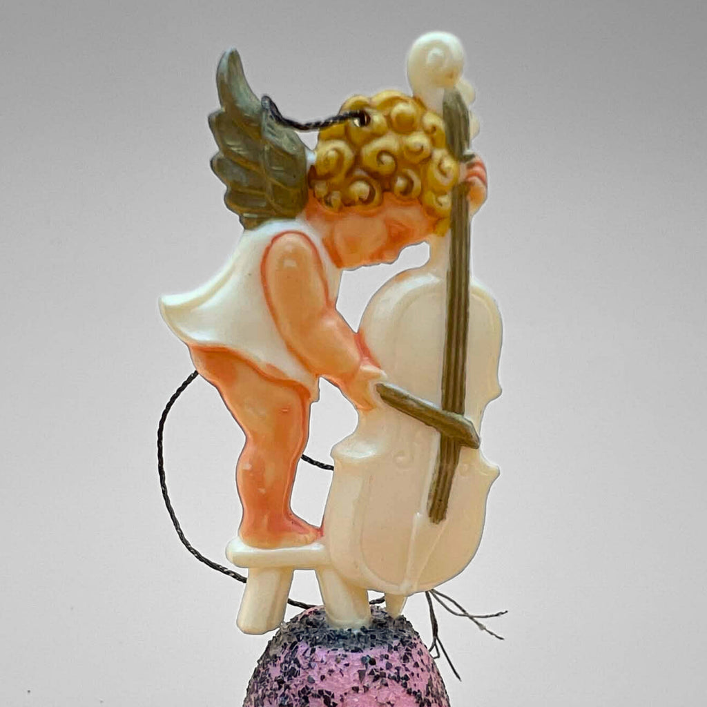 Vintage angel bell ornament with cherub playing cello while standing on a small stool atop a pink glitter bell. Side view highlights sculpted instrument and figure detail. Mid century German Christmas decor collectible