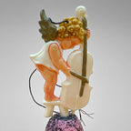 Vintage angel bell ornament with cherub playing cello while standing on a small stool atop a pink glitter bell. Side view highlights sculpted instrument and figure detail. Mid century German Christmas decor collectible