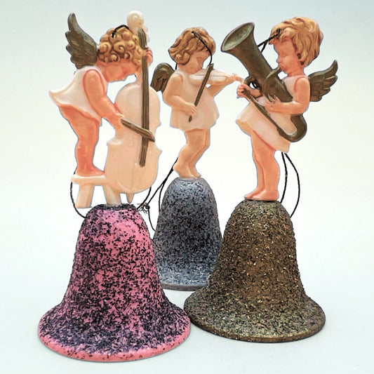 Vintage angel bell ornaments set of three featuring musical cherubs playing cello, violin, and horn instruments. Pink, silver, and gold glitter bell bases shown in front view. Mid century West Germany Christmas decor collectible with hanging strings
