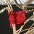 Detail of a worn red paper lighting tag wrapped around the ivory-colored cord of a vintage angel tree topper. Black printing includes decorative lighting wording and the numbers B-997 and E-41423, with tears, creases, and frayed paper edges visible among the cable loops.
