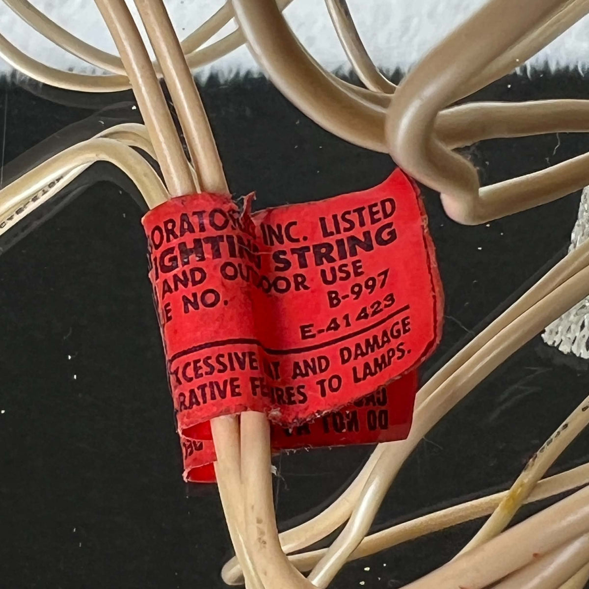Detail of a worn red paper lighting tag wrapped around the ivory-colored cord of a vintage angel tree topper. Black printing includes decorative lighting wording and the numbers B-997 and E-41423, with tears, creases, and frayed paper edges visible among the cable loops.