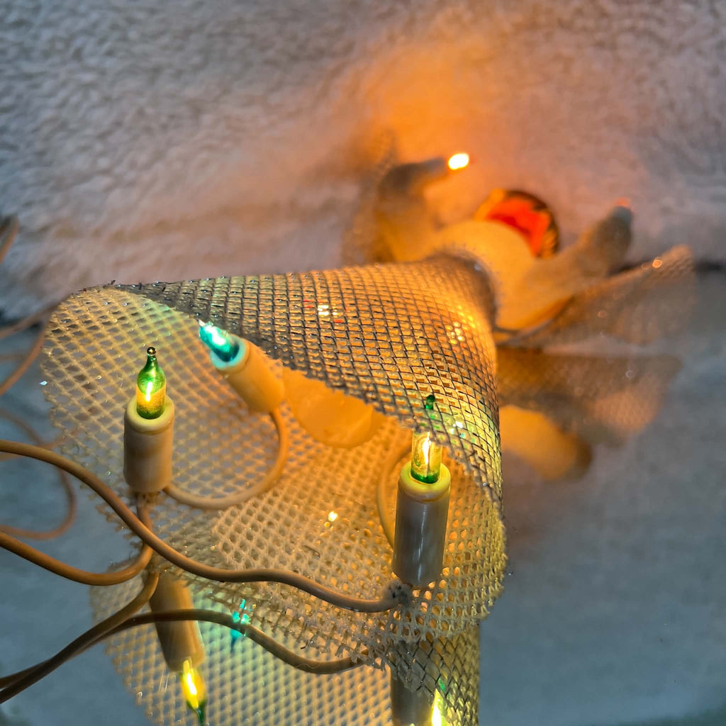 View beneath the illuminated vintage angel topper, showing glowing green and blue bulbs in white sockets inside the silver mesh skirt. Ivory-colored wiring crosses the foreground, while the angel’s face and raised candle light glow warmly in the background.