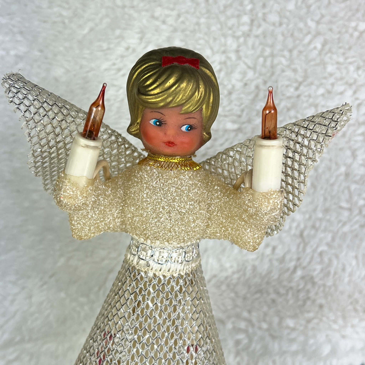 Upper front view of a vintage Christmas angel tree topper against a pale textured background. Gold-colored curls and a red bow frame her painted face, while raised amber candle bulbs, silver mesh wings, a cream textured bodice, and metallic waist trim are visible.