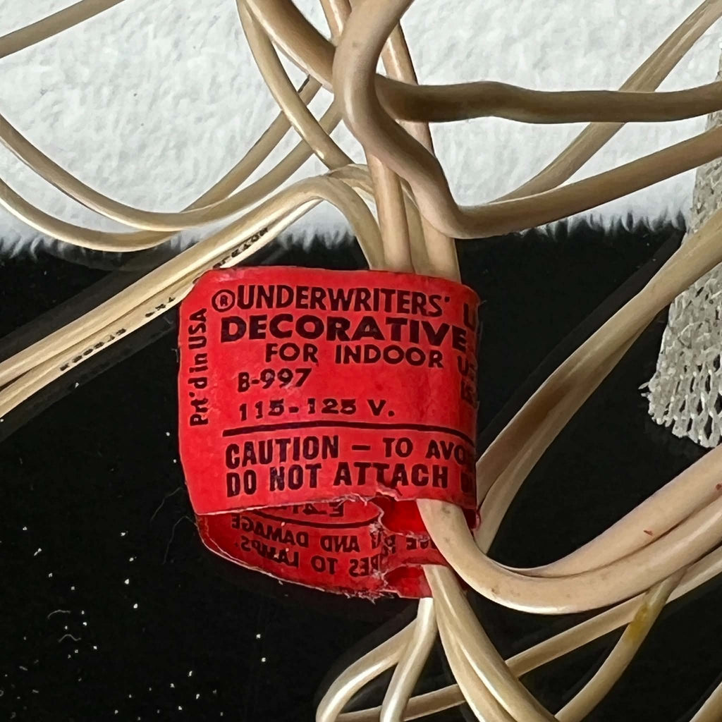 Close view of the red UL lighting label attached to the vintage angel topper’s ivory-colored cord. The tag shows 115–125 V, indoor-use wording, and part of a caution statement, with torn lower edges and creases; the surrounding cord has visible surface marks.