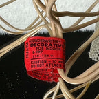 Close view of the red UL lighting label attached to the vintage angel topper’s ivory-colored cord. The tag shows 115–125 V, indoor-use wording, and part of a caution statement, with torn lower edges and creases; the surrounding cord has visible surface marks.