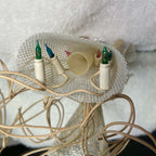 Underside of the unlit vintage angel tree topper, showing its silver mesh skirt, hollow cream mounting tube, colored flame-shaped bulbs, and white sockets. Ivory-colored wiring loops around the skirt opening while the angel rests on a pale textured surface.
