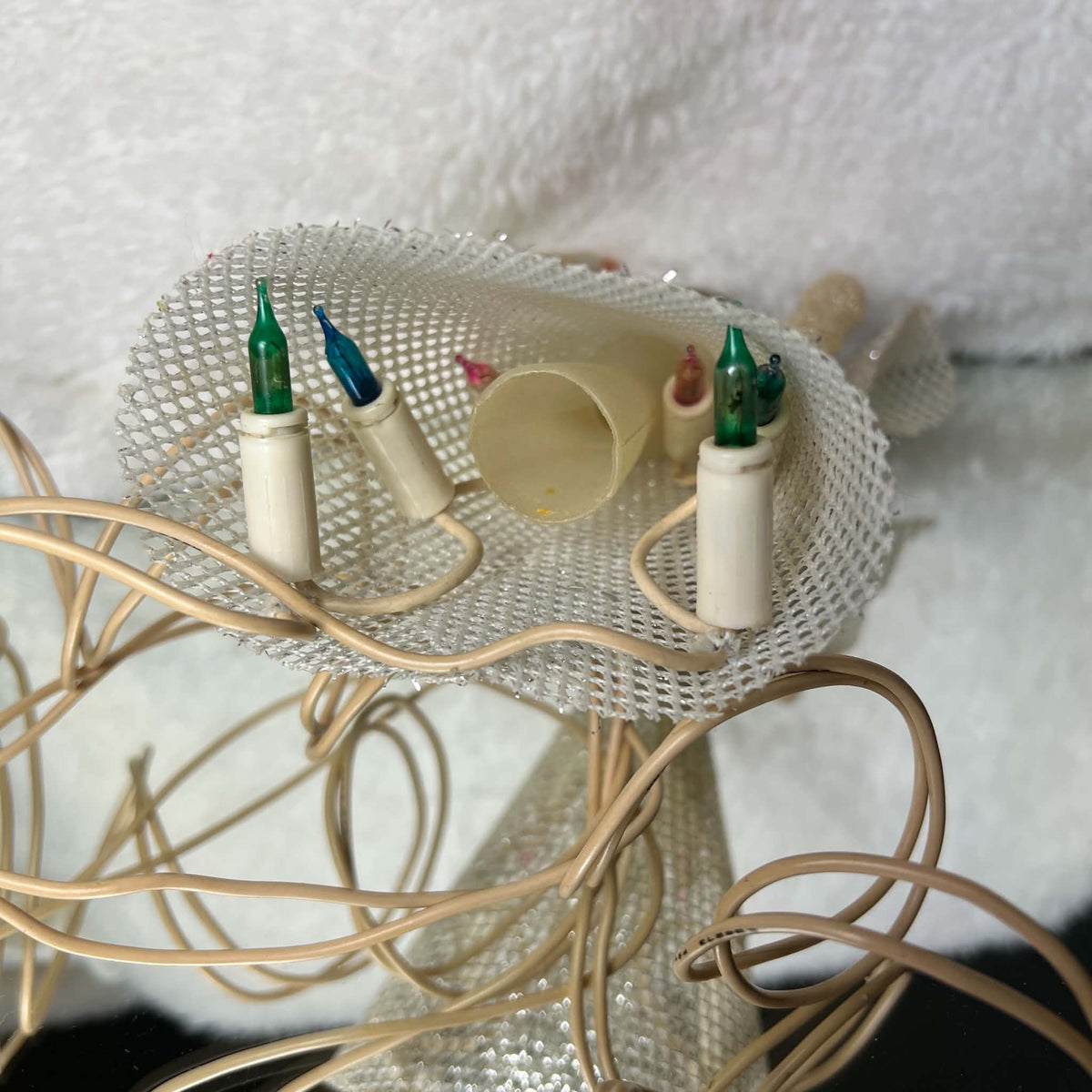 Underside of the unlit vintage angel tree topper, showing its silver mesh skirt, hollow cream mounting tube, colored flame-shaped bulbs, and white sockets. Ivory-colored wiring loops around the skirt opening while the angel rests on a pale textured surface.