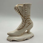 Vintage Atlantic Mold ceramic planter shown in profile as an ivory Victorian high-button boot. Raised round buttons follow the curved front panel, with brown antiquing defining the folds, seams, slender heel and scrolling pedestal base against a neutral background.