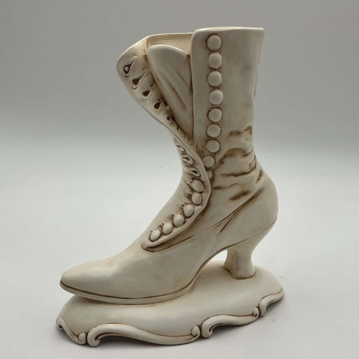 Vintage Atlantic Mold ceramic planter shown in profile as an ivory Victorian high-button boot. Raised round buttons follow the curved front panel, with brown antiquing defining the folds, seams, slender heel and scrolling pedestal base against a neutral background.