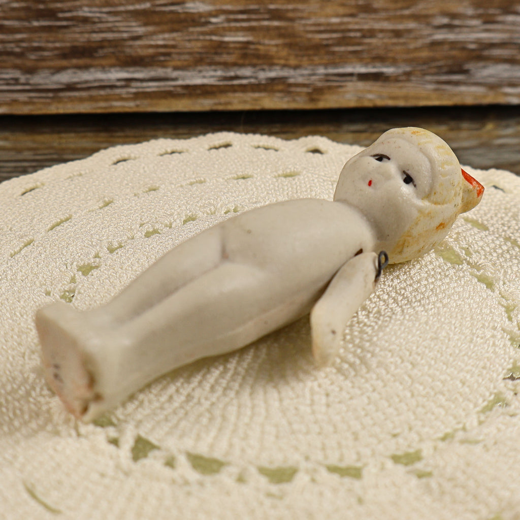 Antique bisque penny doll lying on her back to show the full body, molded bare feet, painted face, red bow, and articulated arms. The close view reveals the doll’s matte ivory surface, scattered age discoloration, worn painted details, and visible joint hardware.
