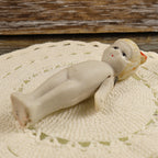 Antique bisque penny doll lying on her back to show the full body, molded bare feet, painted face, red bow, and articulated arms. The close view reveals the doll’s matte ivory surface, scattered age discoloration, worn painted details, and visible joint hardware.