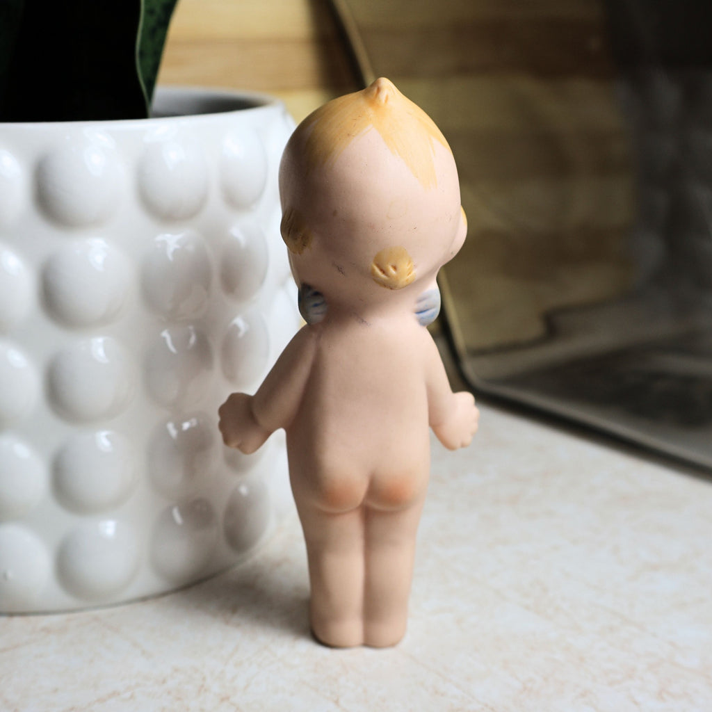 Rear view of the 4.25-inch vintage bisque Kewpie doll showing molded hair detail, painted blond accents, back sculpting, and classic standing pose. This photograph illustrates the overall condition of the collectible from behind.