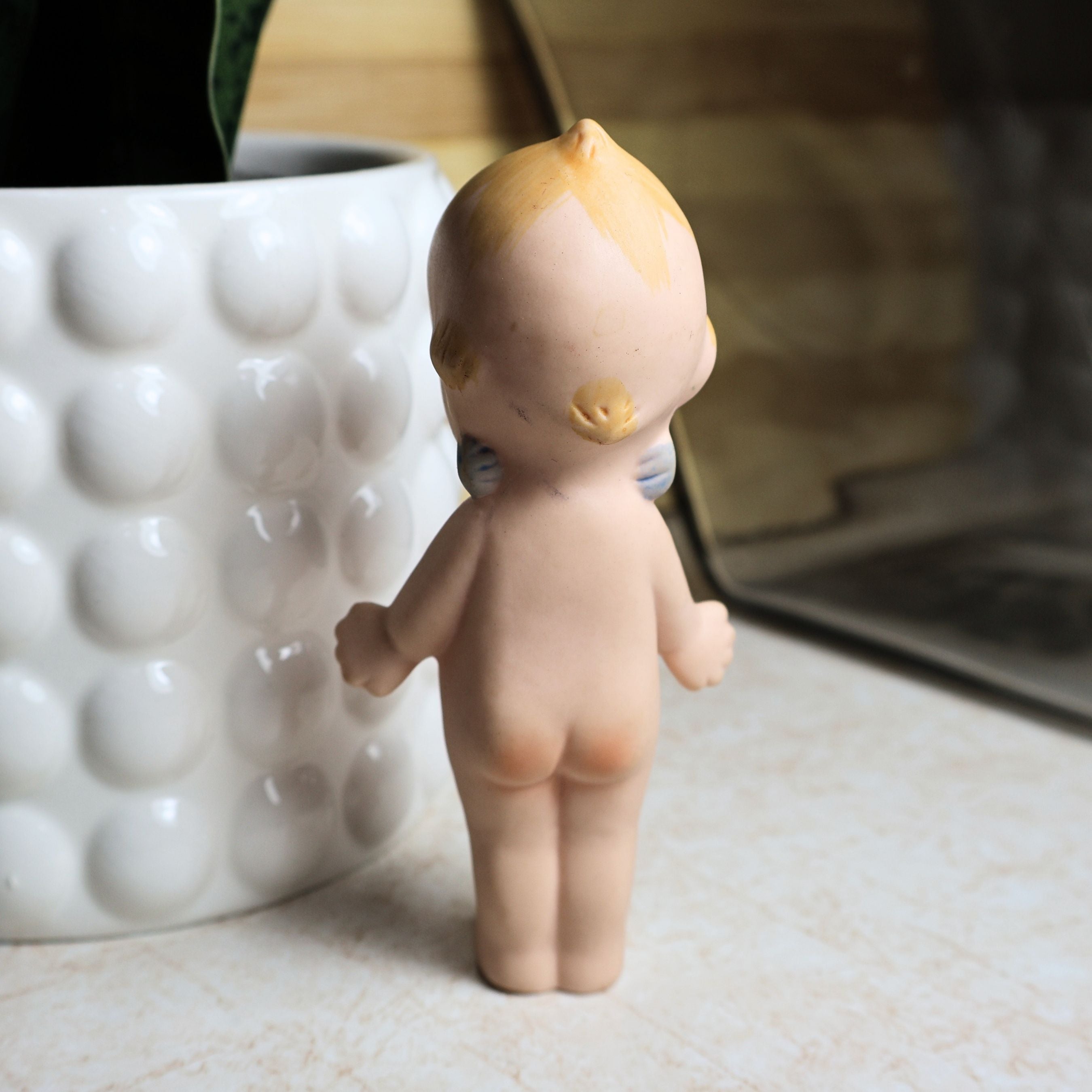 Rear view of the 4.25-inch vintage bisque Kewpie doll showing molded hair detail, painted blond accents, back sculpting, and classic standing pose. This photograph illustrates the overall condition of the collectible from behind.