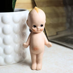 Front view of a vintage 4.25-inch bisque Kewpie doll standing upright with hand-painted facial features, rosy cheeks, blond topknot, and classic side-glancing eyes. The unmarked collectible is photographed against a softly styled neutral background.