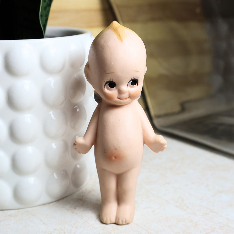 Front view of a vintage 4.25-inch bisque Kewpie doll standing upright with hand-painted facial features, rosy cheeks, blond topknot, and classic side-glancing eyes. The unmarked collectible is photographed against a softly styled neutral background.