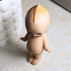Overhead view of the vintage bisque Kewpie doll emphasizing the molded topknot, painted blond hair, expressive downward gaze, and smooth bisque finish while documenting the overall condition from above.
