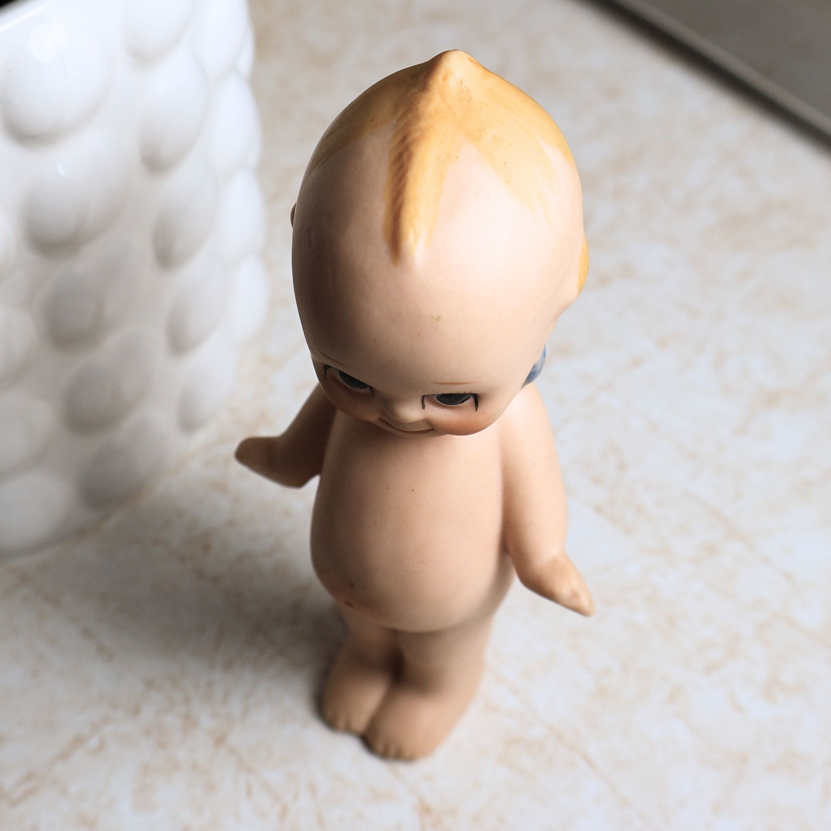 Overhead view of the vintage bisque Kewpie doll emphasizing the molded topknot, painted blond hair, expressive downward gaze, and smooth bisque finish while documenting the overall condition from above.