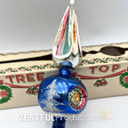 Retro mercury glass Christmas tree topper with blue glass body, silver accents, and colorful finial point displayed in front of original mid century holiday ornament packaging.