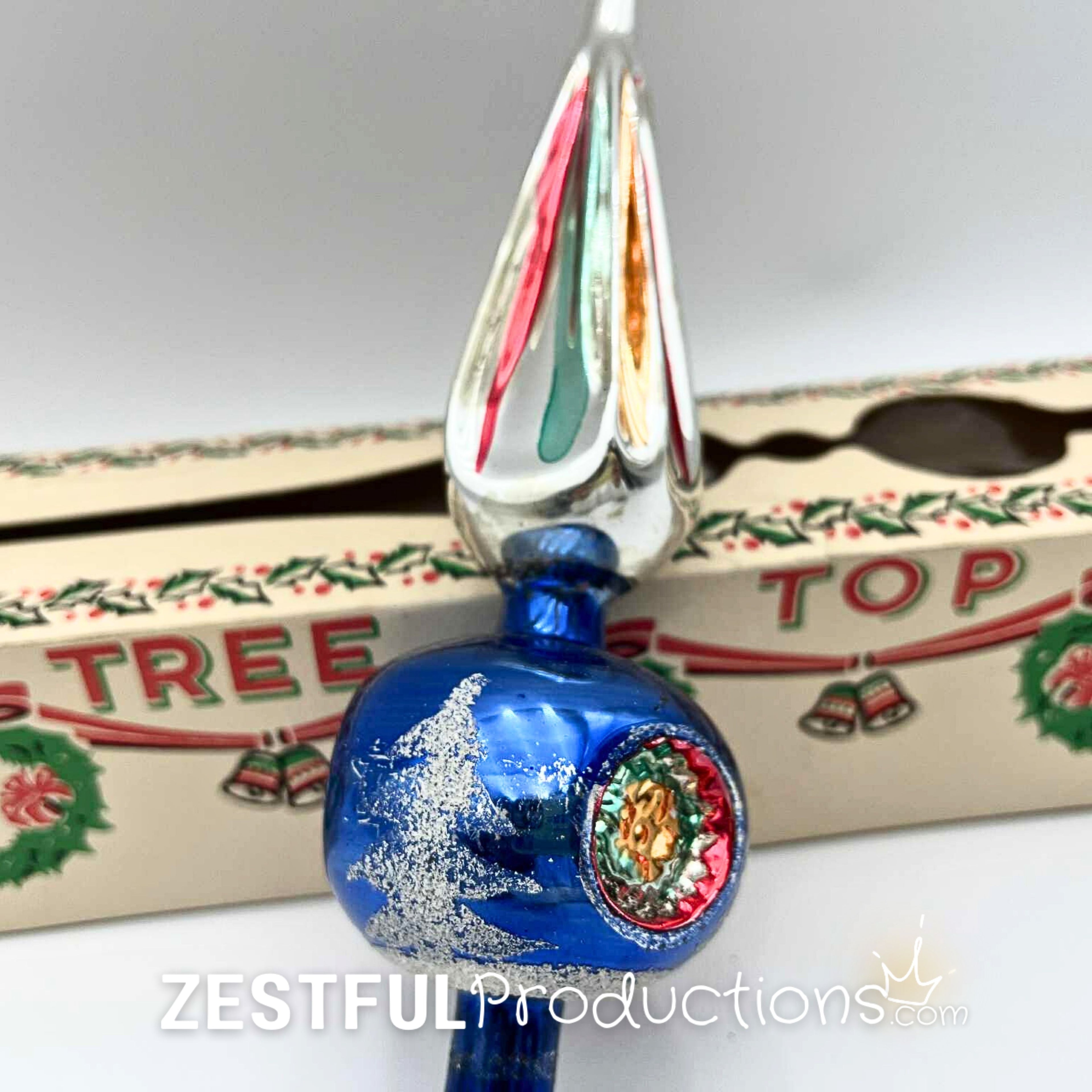 Retro mercury glass Christmas tree topper with blue glass body, silver accents, and colorful finial point displayed in front of original mid century holiday ornament packaging.