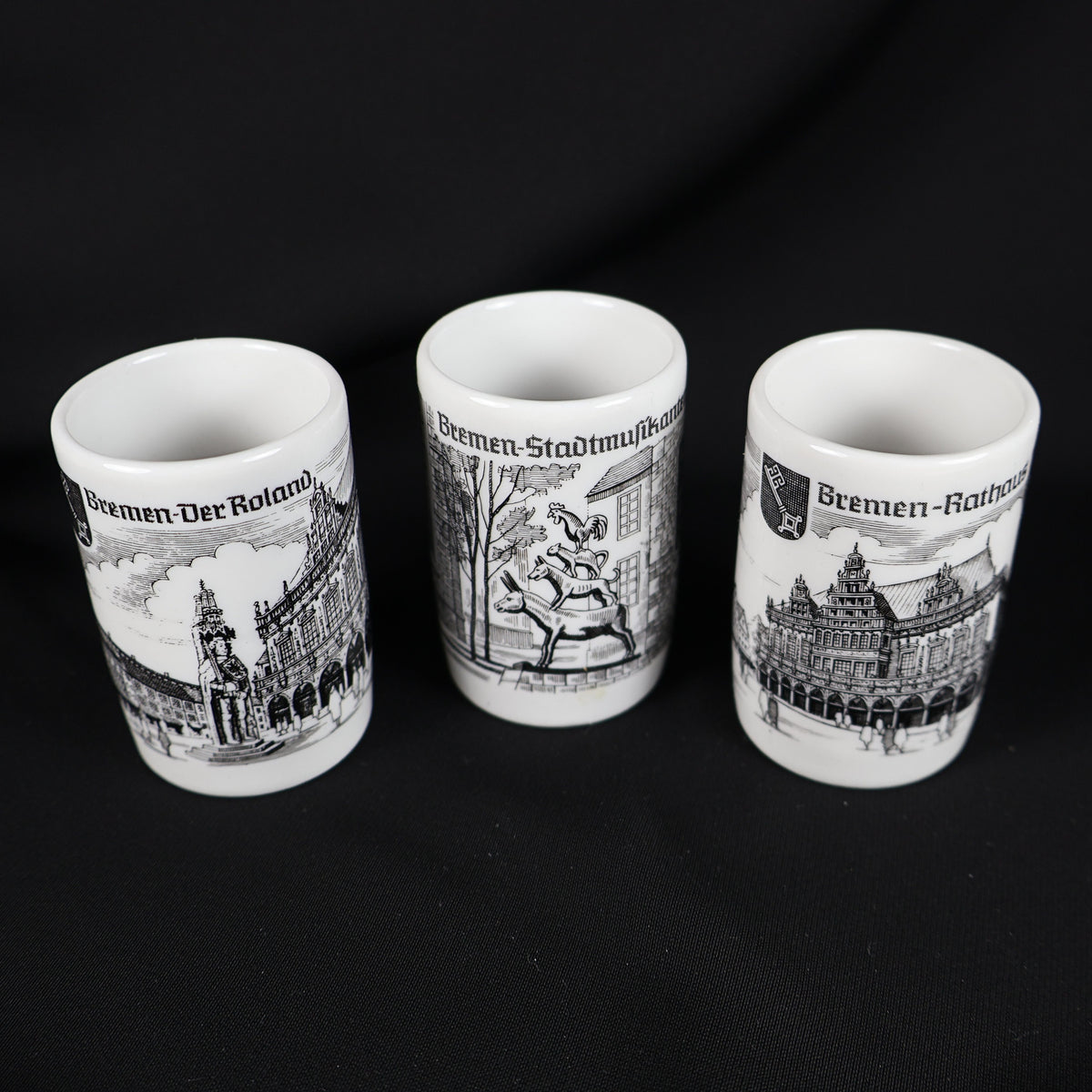 Set of three vintage Bremen Germany porcelain shot glasses arranged against a black background. The white cylindrical cups feature black architectural illustrations labeled Bremen Der Roland, Bremen Stadtmusikanten, and Bremen Rathaus, with their open rims visible.