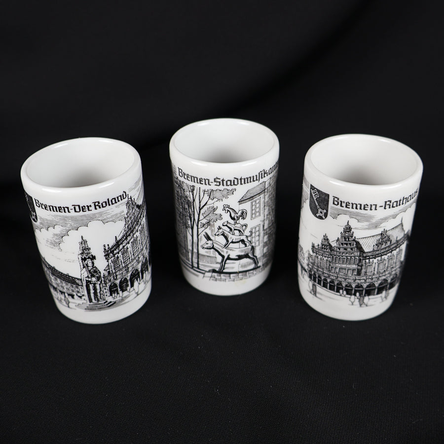 Set of three vintage Bremen Germany porcelain shot glasses arranged against a black background. The white cylindrical cups feature black architectural illustrations labeled Bremen Der Roland, Bremen Stadtmusikanten, and Bremen Rathaus, with their open rims visible.