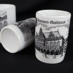 Close view of the vintage Bremen Rathaus porcelain shot glass showing an intricate black drawing of the historic Town Hall beneath its German label. Two coordinating souvenir cups rest behind it, including one placed on its side with an unmarked white base visible.