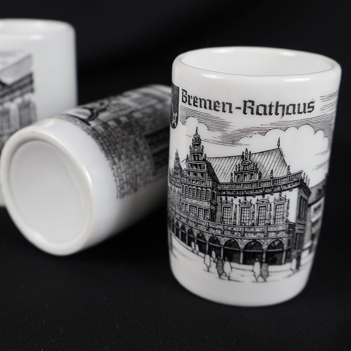 Close view of the vintage Bremen Rathaus porcelain shot glass showing an intricate black drawing of the historic Town Hall beneath its German label. Two coordinating souvenir cups rest behind it, including one placed on its side with an unmarked white base visible.