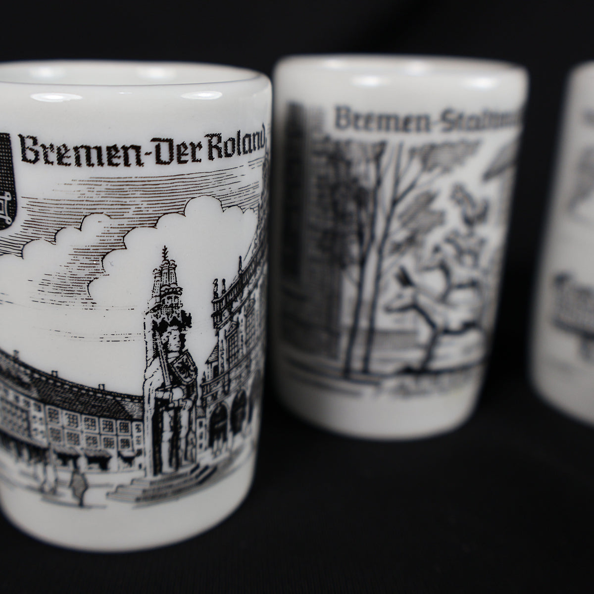Close-up photograph focused on the vintage Bremen Der Roland porcelain shot glass. The black-and-white design shows the standing Roland statue and surrounding marketplace architecture, while the Town Musicians and Rathaus cups appear softly blurred in the background.