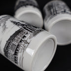 Angled detail photograph of the underside and lower edge of a vintage Bremen Der Roland porcelain souvenir cup. The black marketplace illustration wraps toward the base, while two additional cups appear behind it with plain recessed white bottoms and no visible maker marks.