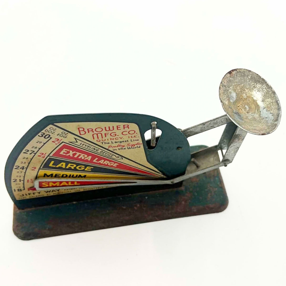 Elevated front view of a Brower-branded Jiffy-Way egg scale on white, showing the colorful sizing chart, open pointer and shallow metal cup. The cup’s interior has mottled discoloration, and the rectangular green base shows extensive brown surface rust.