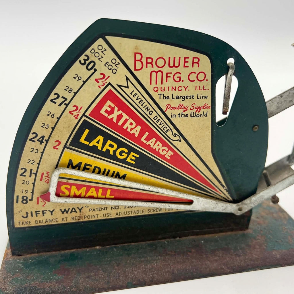 Detail of the Brower Mfg. Co. graphic panel on a Jiffy-Way egg scale, showing Quincy, Illinois lettering, ounce markings and red, yellow and black egg-size bands. An open metal pointer crosses the lower chart above a green-painted base with visible rust.