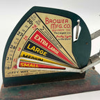 Detail of the Brower Mfg. Co. graphic panel on a Jiffy-Way egg scale, showing Quincy, Illinois lettering, ounce markings and red, yellow and black egg-size bands. An open metal pointer crosses the lower chart above a green-painted base with visible rust.