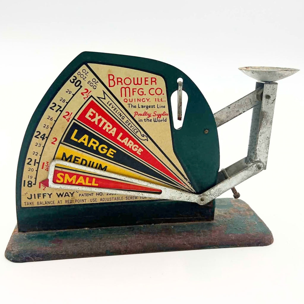 Elevated front view of a Brower-branded Jiffy-Way egg scale on white, showing the colorful sizing chart, open pointer and shallow metal cup. The cup’s interior has mottled discoloration, and the rectangular green base shows extensive brown surface rust.