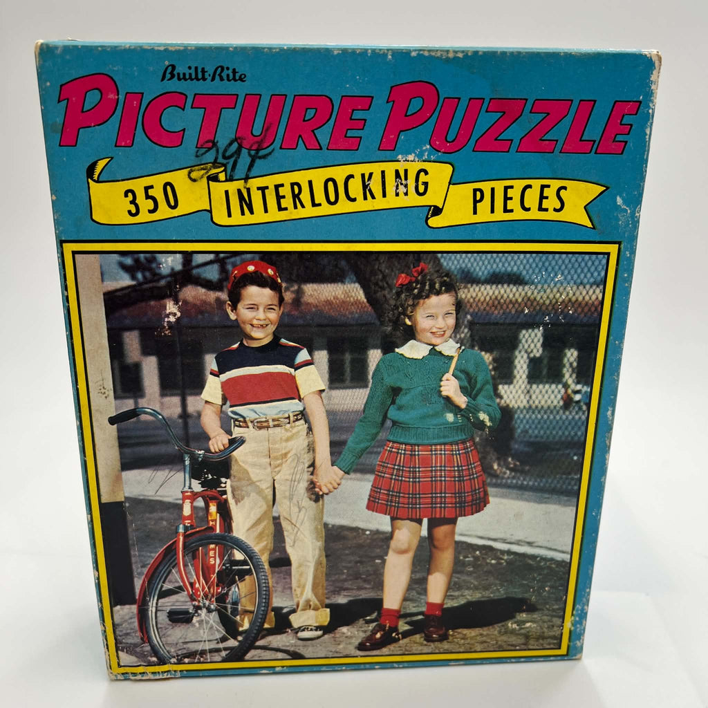 Front of the vintage Built-Rite Ours to Protect picture puzzle box featuring two children holding hands beside a red bicycle. Blue packaging surrounds pink Picture Puzzle lettering and a yellow 350-piece banner. Handwriting and surface wear are visible on the lid.