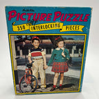 Front of the vintage Built-Rite Ours to Protect picture puzzle box featuring two children holding hands beside a red bicycle. Blue packaging surrounds pink Picture Puzzle lettering and a yellow 350-piece banner. Handwriting and surface wear are visible on the lid.