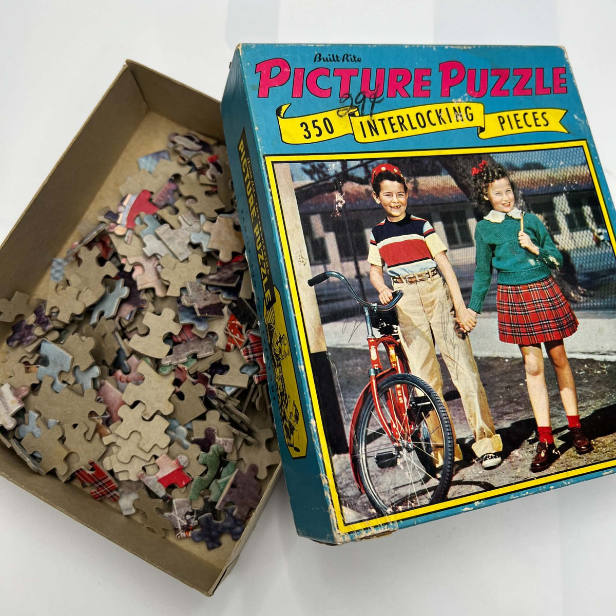 Open vintage Built-Rite Ours to Protect puzzle with loose cardboard pieces beside its illustrated lid. The blue box features pink lettering, a yellow banner, and a photograph of two children holding hands beside a red bicycle. Box edges show visible wear.