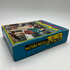 Side view of the vintage Built-Rite puzzle box with a black and yellow Picture Puzzle panel, an illustration of people assembling a puzzle, and the words Family Fun for Everyone. The illustrated lid and visible edge wear appear above the blue side panel.