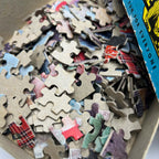 Close-up of loose cardboard pieces from the vintage Built-Rite Ours to Protect picture puzzle. Printed sections show red plaid, green clothing, and parts of a bicycle, while overturned pieces reveal plain cardboard backs and the thickness of the material.