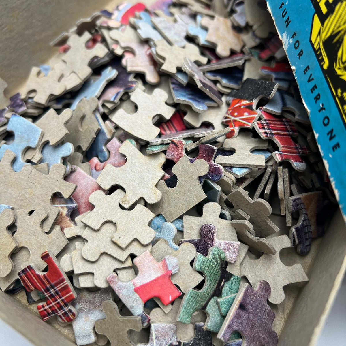 Close-up of loose cardboard pieces from the vintage Built-Rite Ours to Protect picture puzzle. Printed sections show red plaid, green clothing, and parts of a bicycle, while overturned pieces reveal plain cardboard backs and the thickness of the material.