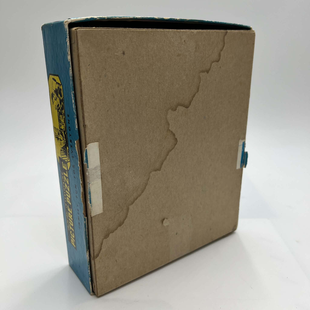 Underside of the original vintage Built-Rite Ours to Protect puzzle box, showing the plain brown cardboard base, visible water damage, and tape remnants along the sides. The worn blue lid surrounds the base, with part of its yellow and black side graphic visible.