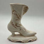 Opposite side of a vintage ivory ceramic Victorian boot planter, showing its tall curved shaft, pointed toe, narrow heel and ornamental pedestal. Hand-applied brown accents settle into the sculpted creases and edges, creating an intentionally antiqued appearance.