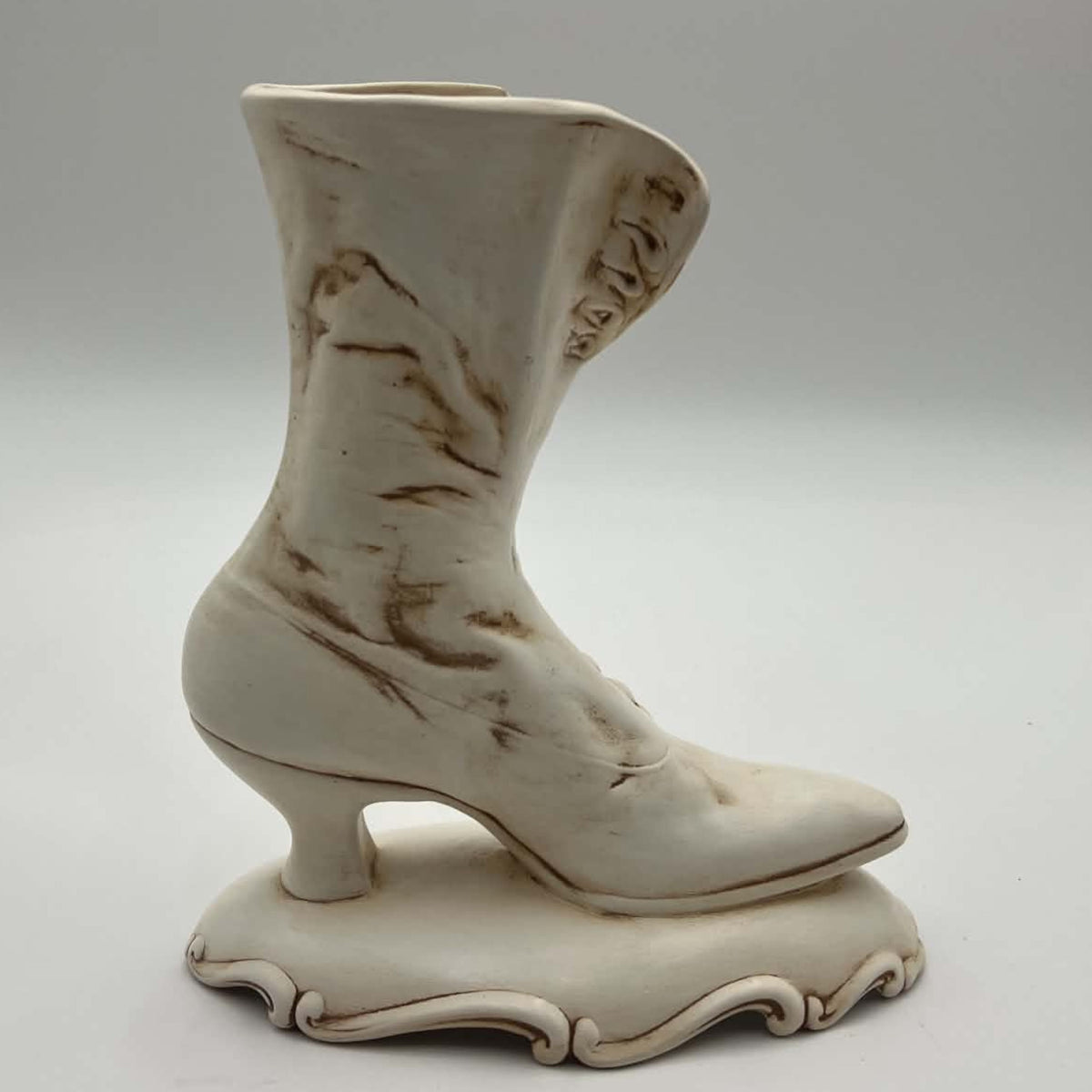 Opposite side of a vintage ivory ceramic Victorian boot planter, showing its tall curved shaft, pointed toe, narrow heel and ornamental pedestal. Hand-applied brown accents settle into the sculpted creases and edges, creating an intentionally antiqued appearance.