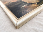 Close-up of a cream-finished wood frame corner around the dark edge of a portrait print. The miter joint, small chips, scuffs, darker exposed areas, and a dark oval mark on the outer edge are visible against the pale woven fabric underneath.