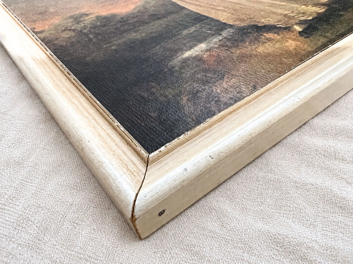 Close-up of a cream-finished wood frame corner around the dark edge of a portrait print. The miter joint, small chips, scuffs, darker exposed areas, and a dark oval mark on the outer edge are visible against the pale woven fabric underneath.