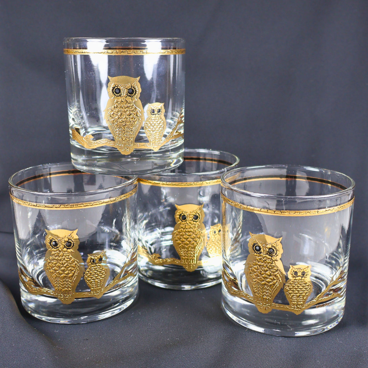 Set of four vintage Culver Ltd. clear rocks glasses arranged against a dark background, each decorated with two textured gold owls perched on a branch. Gold bands circle the rims, while black eye accents add expression to the mid-century barware design.