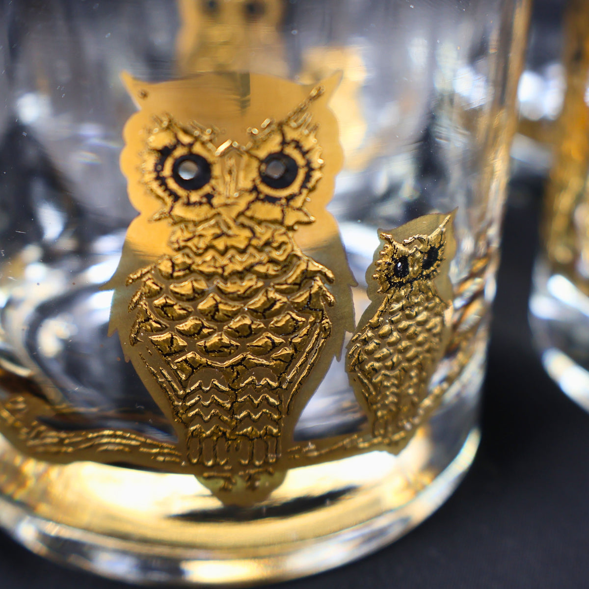 Close angled view through a vintage Culver cocktail glass, focusing on a textured gold owl with dark eyes and a second smaller owl behind it. Reflections in the heavy clear glass emphasize the dimensional decoration and substantial lowball form.
