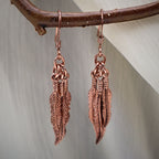 Close up of vintage feather dangle earrings in copper tone finish hanging on natural branch, detailed western boho jewelry design with layered feather charms, lightweight statement earrings for women, nature inspired accessory perfect for festival wear, rustic bohemian style jewelry gift for her