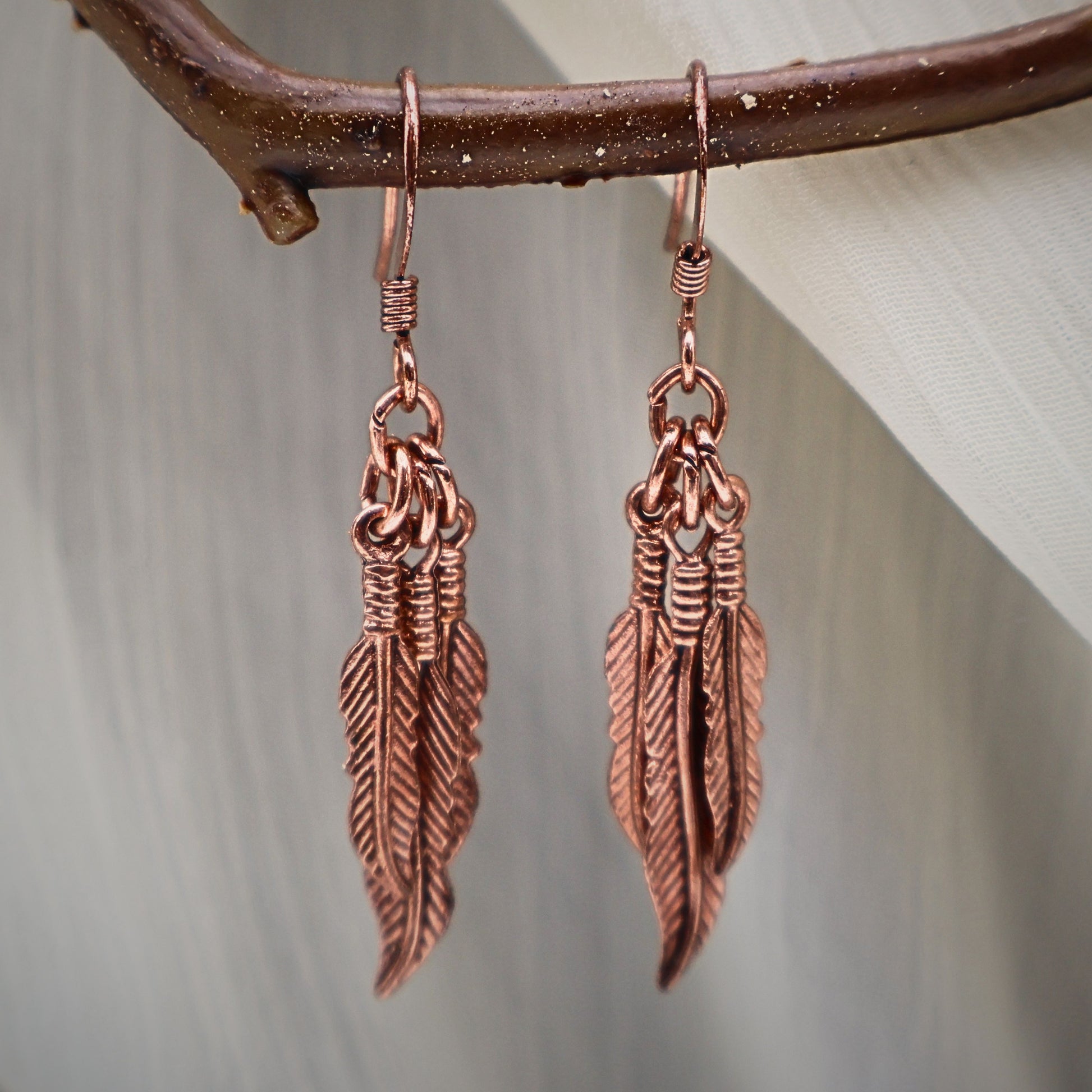 Close up of vintage feather dangle earrings in copper tone finish hanging on natural branch, detailed western boho jewelry design with layered feather charms, lightweight statement earrings for women, nature inspired accessory perfect for festival wear, rustic bohemian style jewelry gift for her
