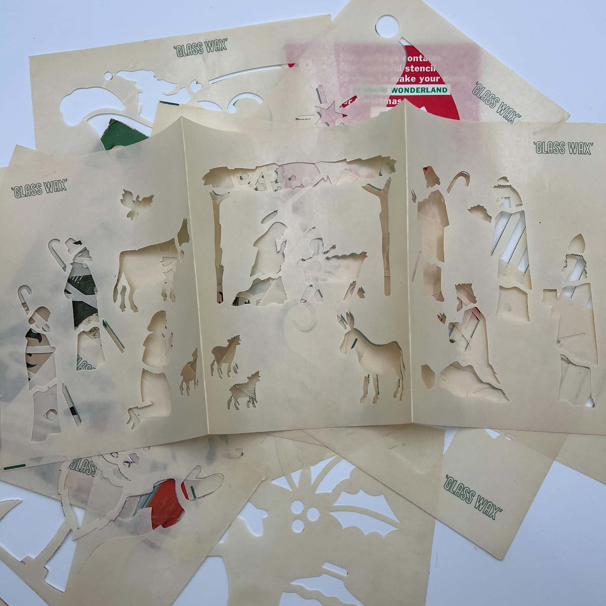 An unfolded vintage Glass Wax Nativity stencil rests across overlapping cream paper stencil sheets. Cutout figures include the manger scene, shepherds, animals, and visitors, with green Glass Wax printing, fold lines, and parts of the original envelopes visible.