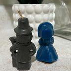Back view of two vintage Gurley Thanksgiving candles showing the charcoal wax clothing and long gun on the male pilgrim and deep blue molded bonnet and dress on the female figure. Both candles display surface wear and marks consistent with age and storage.